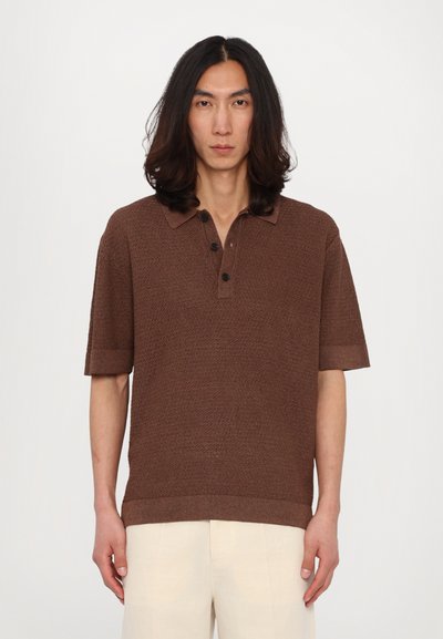 Bruine gebreide poloshirt met een gestructureerd patroon, drie zwarte knopen en korte mouwen, gecombineerd met lichtkleurige linnen broek.