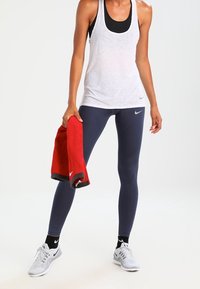 Kobieta w białym topie na ramiączkach i czarnym biustonoszu sportowym, ubrana w granatowe legginsy i białe sneakersy, trzyma w jednej ręce czerwoną ręcznik Nike.