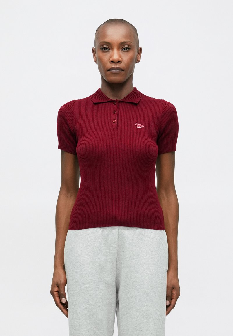 Sieviete, tērpta pieguļošā burgundijas krāsas īsajās piedurknēs, ribota polo krekla ar trim pogām un mazu logo uz krūtīm, savienota ar gaiši pelēkiem biksēm.