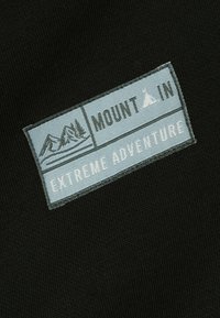 Vêtement noir présentant une pièce rectangulaire en tissu bleu clair. La pièce affiche "MOUNT" et "EXTREME ADVENTURE" imprimés en noir, accompagnés de graphismes de montagnes.