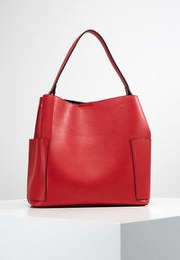 Červená kožená taška přes rameno se strukturovaným tvarem, vybavená dvěma postranními kapsami a jedním uchopovacím držadlem. Hladký povrch, minimalistický design.