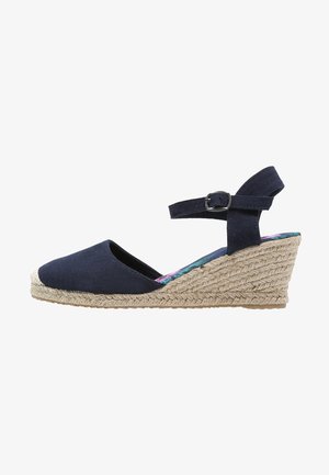 Sandale compensée bleu marine avec bout fermé, bride à la cheville avec boucle et motif en corde tressée sur un talon de hauteur moyenne.