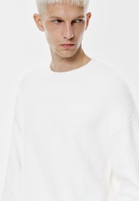 Biały sweter wykonany z miękkiego, teksturowanego materiału z okrągłym dekoltem. Posiada opadające ramiona i luźny krój.