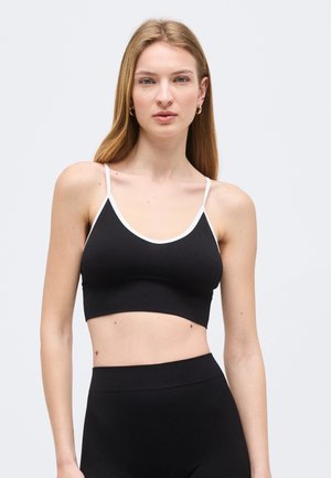 Ung kvinde med langt blondt hår iført en sort cropped tanktop med hvid kant og sorte højtragtede bukser, stående mod en lys baggrund.