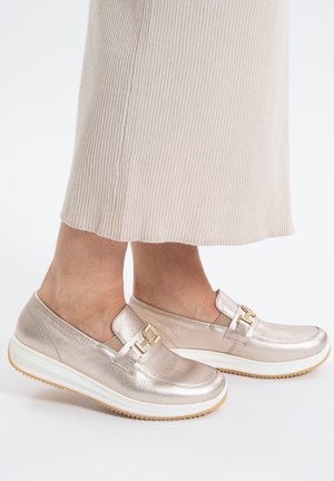 Feet wearing metallic slip-on loafers with gold buckle detail and white thick rubber soles, paired with beige ribbed wide-leg pants.