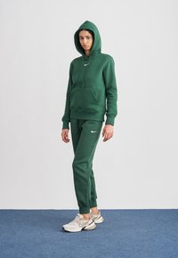 Zöld pamut kapucnis pulóver első zsebbel, hozzáillő melegítőnadrággal párosítva. A felsőrészen és a nadrágon fehér Nike-logó található. A modell világos sportcipőt visel.