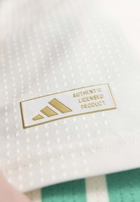 Weißer Stoff mit strukturierter Musterung, einem auffälligen gold-weißen rechteckigen Etikett mit der Aufschrift "ECHTES LIZENZIERTES PRODUKT" und drei Streifen.