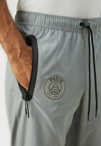 Gray athletic joggers with an elastic waistband, black zipper pocket, and a circular logo featuring "PARIS SAINT-GERMAIN" in gray.