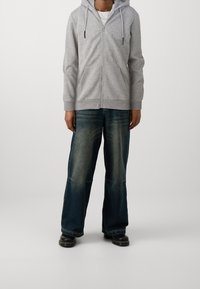 Un sweat à capuche zippé gris avec des cordons, des poches avant, associé à un jean large en denim foncé, avec un lavage décoloré et des chaussures noires.