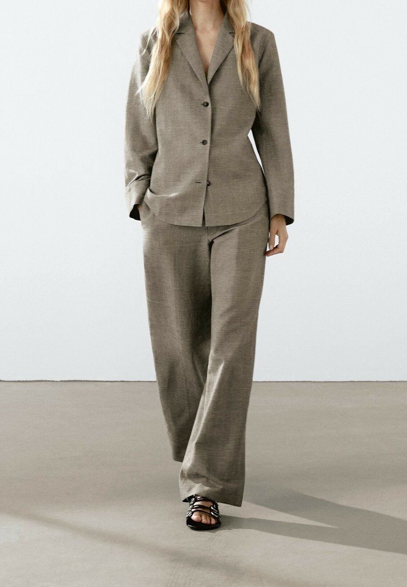 Femme portant un blazer taupe ample et un pantalon large assorti, marchant vers l'avant les mains dans les poches, portant des sandales noires.