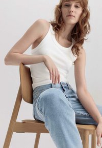 Witte geribbelde tanktop met een wijde halslijn, gecombineerd met lichtblauwe rechte jeans, zittend op een houten stoel tegen een effen achtergrond.