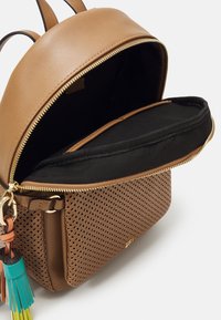 Sac à dos en cuir marron avec poche avant perforée, fermeture éclair dorée partiellement ouverte dévoilant une housse pour ordinateur portable noire et matelassée à l'intérieur, et porte-clés pompon coloré.