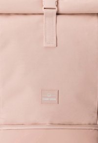 Svetloružový batoh zo štruktúrovanej textílie, s preklopeným vrchným dielom, zapínaním na remienok a malou logovou značkou.