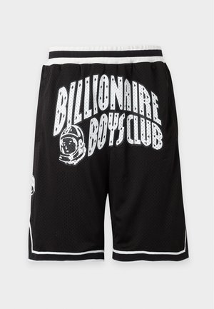 Mladý muž stojí s rukami v vreckách, oblečený v bielom tričku Billionaire Boys Club a čiernych šortkách s rovnakým logom a čiernych teniskách.