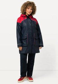 Veste imperméable marine avec des accents rouges, capuche ajustable, deux poches avant et cordons de serrage à motifs ; associée à un pantalon sombre et des chaussures à plateforme.