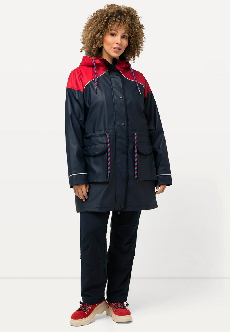Veste imperméable marine avec des accents rouges, capuche ajustable, deux poches avant et cordons de serrage à motifs ; associée à un pantalon sombre et des chaussures à plateforme.