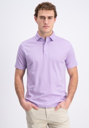 Jonge man staat met handen in de zakken, draagt een lichtpaars polo met korte mouwen en beige broek tegen een witte achtergrond.