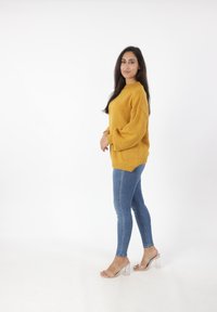 Maglione oversize giallo senape con dettagli a coste, abbinato a jeans in denim blu aderenti e sandali con tacco a blocco trasparente.