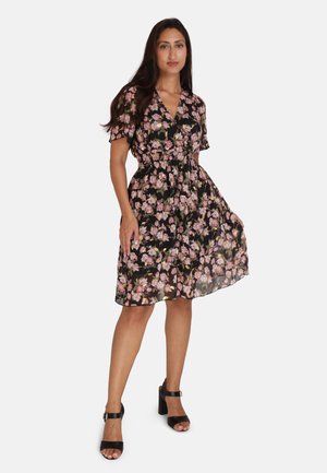 Zwarte bloemenjurk met korte mouwen, elastische taille en vloeiende rok met rozen- en groene bloemenpatronen. Gekozen met zwarte hoge sandalen.