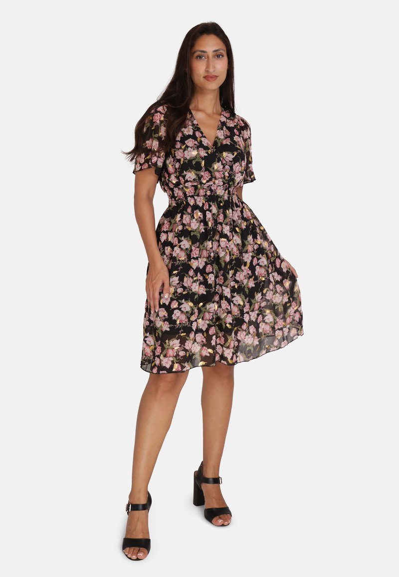 Vestido negro de flores con mangas cortas, cintura elástica y falda fluida con patrones de flores rosas y verdes. Se lleva con sandalias de tacón negras.