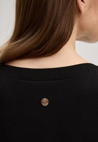 Μαύρο πουλόβερ με πλεκτή λαιμόκοψη και κυκλικό χρυσό ταμπελάκι με σχέδιο λουλούδι στο πίσω μέρος. Ορατή απαλή πλεκτή υφή.