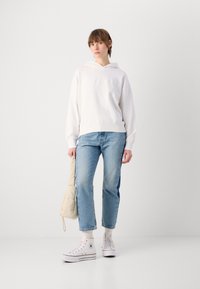 Felpa bianca con cappuccio e logo in rilievo, abbinata a jeans a gamba dritta di colore azzurro chiaro e sneakers bianche. È in mano una borsa imbottita beige.