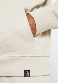 Beige sweatshirt med känguruficka, ribbad muddar och en svart logotyp på fållen. Mjuk tyg med en jämn textur.