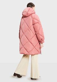 Abrigo acolchado rosa con capucha, con un patrón de diamantes y puños elásticos. Usado sobre pantalones anchos color crema.