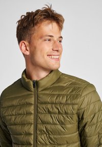 Oliven grøn, let quiltet pufferjakke med højt krave og frontlynlås. Har horisontal syning og glat tekstur.