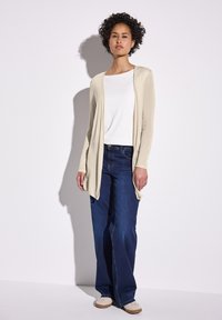 Beige pitkä neulecardigan valkoisen topin päällä, yhdistettynä sinisiin farkkuihin. Cardiganissa on avonainen etuosa ja pehmeä pinta. Beiget kengät viimeistelevät asun.