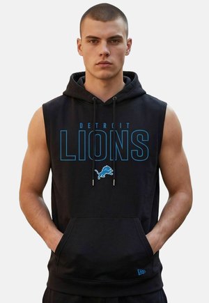 Młody mężczyzna z krótkimi włosami, ubrany w czarną kamizelkę z kapturem z logo Detroit Lions i napisem, ręce w przedniej kieszeni.