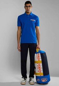 Camisa polo azul de manga corta con logo bordado, combinada con pantalones oscuros y zapatillas blancas. Sosteniendo un gran bolso de viaje multicolor.
