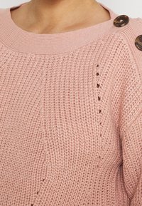 Pull tricoté rose clair avec un col rond côtelé, des boutons décoratifs sur l'épaule et des motifs de couture texturés.