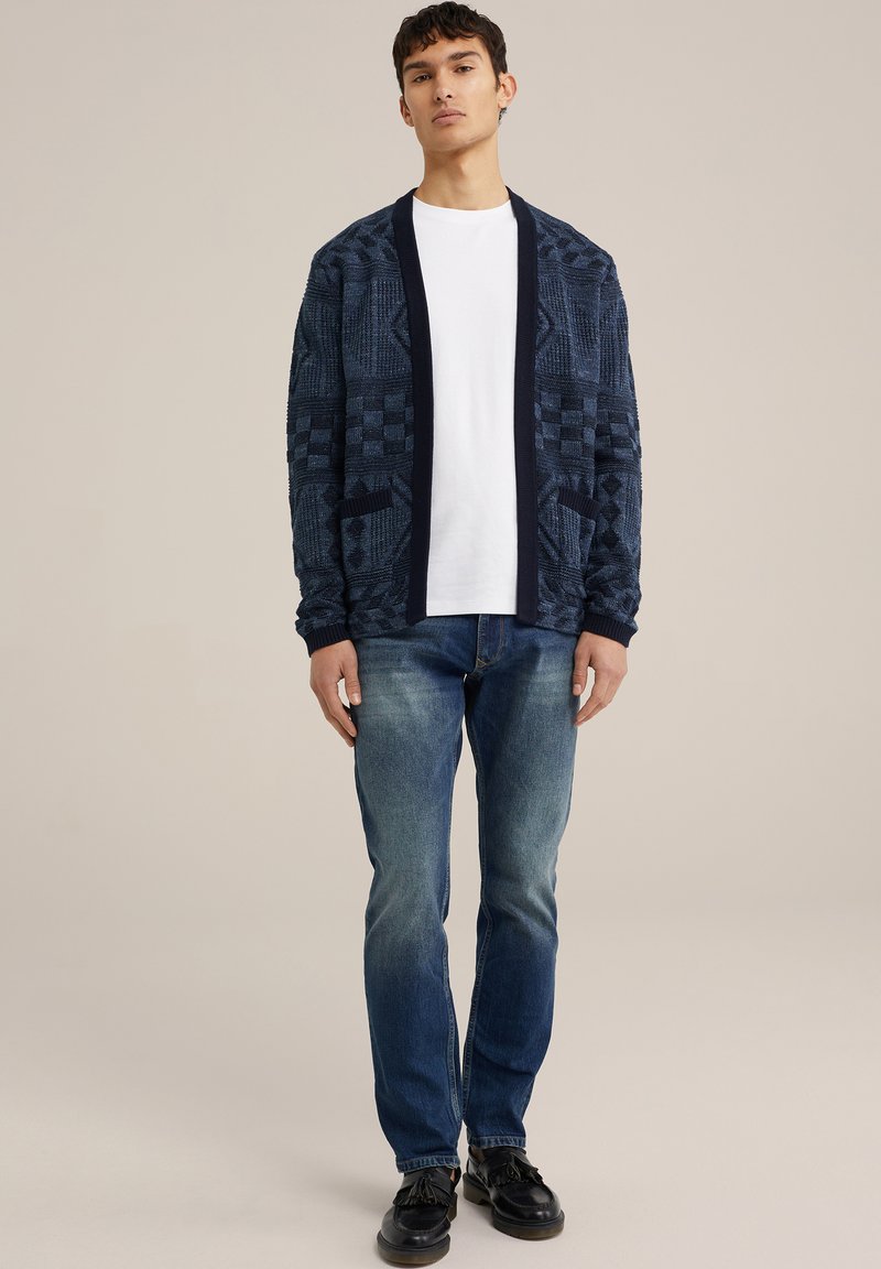 Maritiem gemusterde cardigan met een sjaalkraag, twee voorzakken, gedragen over een wit shirt; gecombineerd met lichtblauwe jeans en zwarte schoenen.
