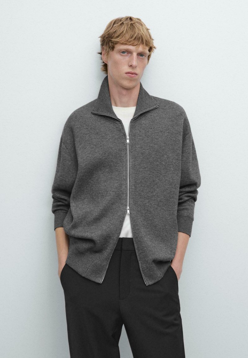 Maglione grigio con zip, colletto alto, vestibilità rilassata e polsini a coste. Indossato sopra una camicia bianca, abbinato a pantaloni neri.
