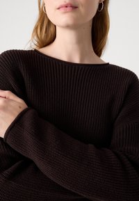 Femme portant un pull en tricot côtelé marron foncé, les bras croisés et des boucles d'oreilles argentées en forme d’anneau, visible du cou jusqu’au milieu du torse.