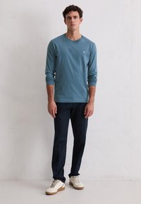 Jasnoniebieska koszula z długim rękawem i okrągłym dekoltem, zestawiona z granatowymi spodniami i białymi trampkami z teksturowaną podeszwą, stojąca na tle jednolitej ściany.