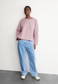 Gestreiftes langärmliges Hemd in Mauve und Weiß, kombiniert mit hellblauen, lässig sitzenden Jeans. Das Model steht vor einer weißen Wand und trägt Sneakers.