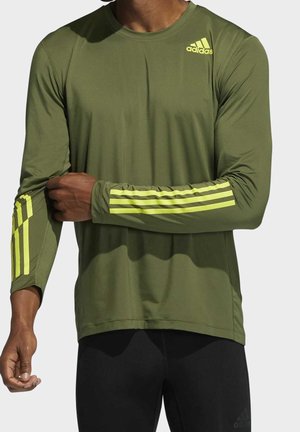 Homme portant un maillot de sport Adidas vert olive à manches longues, avec des bandes jaune néon sur les manches et un logo sur la poitrine, assorti à un pantalon noir.