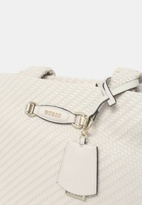 Borsa a mano intrecciata bianca con un design elegante, dettagli in ecopelle, una placca con il logo dorato "Guess" e un'etichetta staccabile. Presenta un materiale testurizzato.