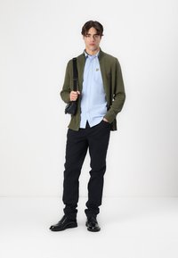 Grønn strikket cardigan, lyseblå skjorte, svarte bukser og svarte sko. Antrekket har en liten logo på cardiganen og en crossbody-veske.