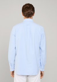 Jasnoniebieska bawełniana koszula z długimi rękawami, z guzikami przy mankietach oraz plisą z tyłu. Posiada kołnierzyk i zaokrąglony dół. Prosty design.