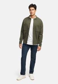 Grön stickad jacka med två bröstfickor, knappstängning, bärs över en vit t-shirt, ihop med mörkblå jeans och vita sneakers.