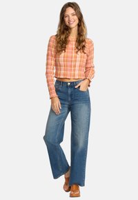 Jeune femme souriante, portant un haut court à manches longues à carreaux orange, un jean large bleu et des sandales à plateforme marron sur fond blanc.