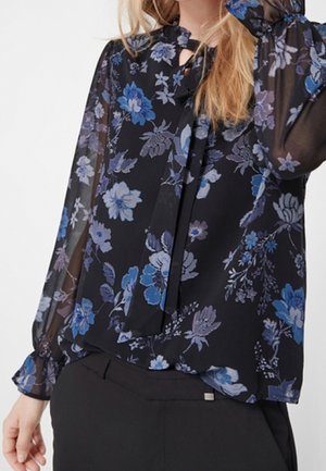 Mujer con blusa negra transparente con estampado floral azul y morado y puños con volantes, combinada con pantalones negros.