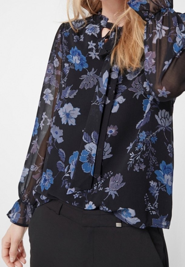 Femme portant un chemisier noir transparent à imprimé floral bleu et violet avec des poignets à volants, associé à un pantalon noir.