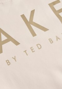 Heleda beež kangas, millel on suured kullavärvi tähed, mis ütlevad "AK E BY TED BAKER." Sile tekstuur ja minimaalsed disainielemendid.