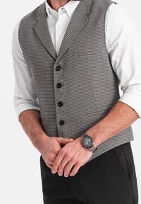 Grå mönstrad väst med en figurnära design, fem svarta knappar och två sidofickor, bärs över en vit skjorta med uppvikta ärmar.