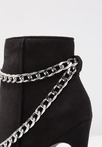 Bottine en daim noir présentant une forme effilée, un accent de chaîne argentée enroulée autour de la bottine, et un talon haut.