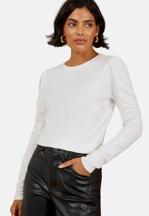 Donna con capelli corti e scuri che indossa una maglietta bianca a maniche lunghe e pantaloni di pelle neri, con una mano in tasca, che guarda di lato.